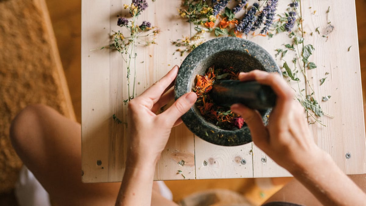 Hands using a mortar and pestle to grind colorful dried herbs on a wooden table.
